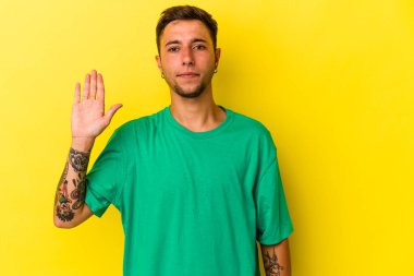 Sarı arka planda izole edilmiş dövmeleri olan beyaz bir genç adam gülümseyerek beş numarayı parmaklarıyla gösteriyor..