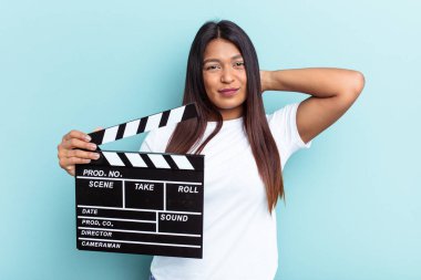 Genç Venezuelalı bir kadın mavi arka planda izole edilmiş bir klempon tutuyor. Kafasının arkasına dokunuyor, düşünüyor ve bir seçim yapıyor..