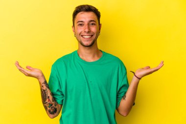 Sarı arka planda dövmeleri olan genç beyaz adam kollarıyla ölçek çiziyor, mutlu ve kendinden emin hissediyor..