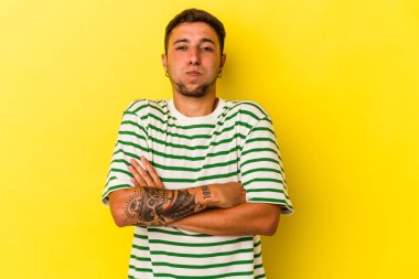 Sarı arka planda izole edilmiş dövmeleri olan beyaz genç bir adam yanakları patlatıyor, yorgun bir ifade takınıyor. Yüz ifadesi kavramı.