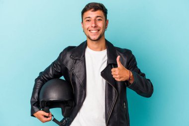 Mavi arka planda kask tutan genç bisikletçi gülümsüyor ve başparmağını kaldırıyor.