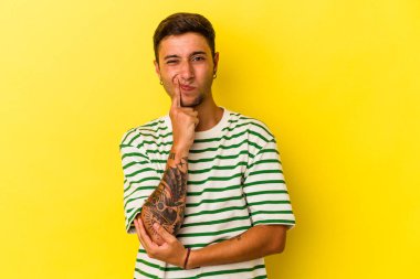 Sarı arka planda izole edilmiş beyaz dövmeli genç adam alaycı bir ifadeyle kameraya bakıyor..