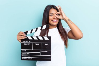 Genç Venezuelalı bir kadın mavi arka planda izole edilmiş bir klempon tutuyor. Heyecanlı. Göze göz işareti yapıyor..