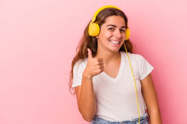 Pembe arka planda izole edilmiş müzik dinleyen beyaz bir kadın gülümsüyor ve başparmağını kaldırıyor.