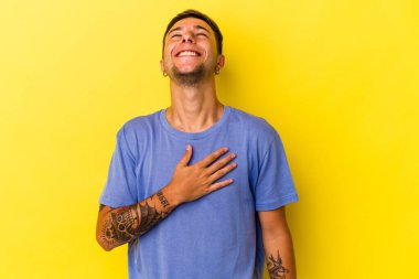 Sarı arka planda izole edilmiş dövmeleri olan beyaz bir genç elini göğsünde tutarak kahkahalar atıyor..