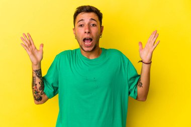 Sarı arka planda izole edilmiş dövmeleri olan genç beyaz bir adam zaferi ya da başarıyı kutluyor, şaşırıyor ve şok oluyor..