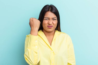 Mavi arka planda izole edilmiş genç Venezuelalı kadın yumruğunu kameraya gösteriyor, agresif yüz ifadesi..