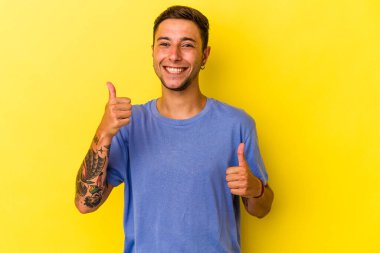 Sarı arka planda izole edilmiş beyaz dövmeli genç bir adam. Baş parmağını kaldırıyor, gülümsüyor ve kendinden emin..