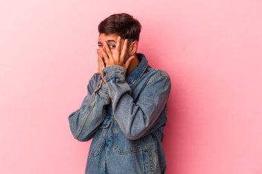 Sarı arka planda izole edilmiş dövmeleri olan beyaz genç bir adam parmaklarının arasından korkmuş ve gergin bir şekilde göz kırpıyor..