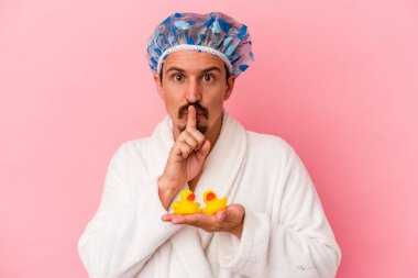 Genç beyaz adam duşa pembe arka planda izole edilmiş plastik ördeklerle giriyor ya sır saklıyor ya da sessizlik istiyor..