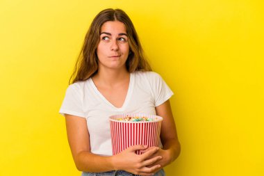 Sarı arka planda izole edilmiş patlamış mısırları tutan genç beyaz kadın kafası karışmış ve kendinden emin değil..