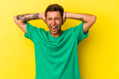 Sarı arka planda izole edilmiş dövmeleri olan genç beyaz bir adam kulaklarını elleriyle kapatıyor ve çok fazla ses duymamaya çalışıyor..