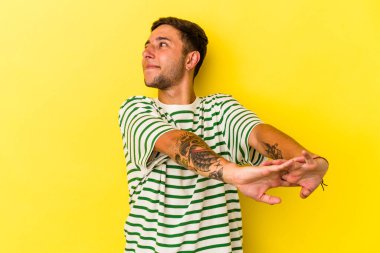 Sarı arka planda izole edilmiş, kollarını esneten, rahat bir pozisyonda dövmeleri olan genç beyaz bir adam..