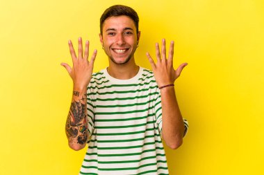 Sarı arka planda izole edilmiş dövmeleri olan genç beyaz bir adam 10 numarayı elleriyle gösteriyor..