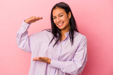 Pembe arka planda izole edilmiş genç Latin kadın işaret parmaklarıyla küçük bir şey tutuyor, gülümsüyor ve kendinden emin..