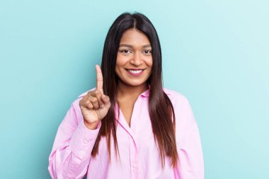 Genç Venezuelalı bir kadın mavi arka planda izole edilmiş. Parmağıyla bir numarayı gösteriyor..