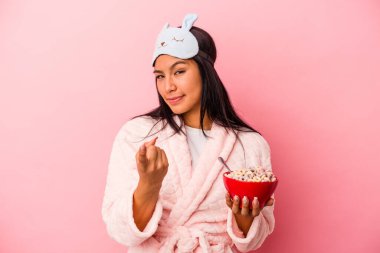 Pembe arka planda bir kase mısır gevreği tutan, pijama giyen genç Latin kadın sanki seni davet ediyormuş gibi seni işaret ediyor..