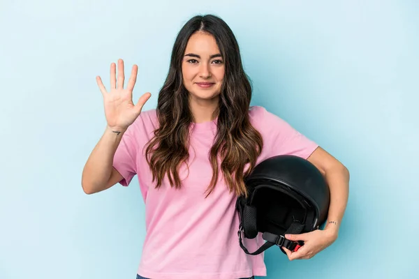 Young caucasian woman holding a motor bike helmet isolated on blue background smiling cheerful showing number five with fingers.