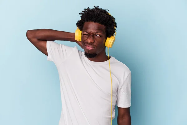 Young african american man listening to music isolated on blue background touching back of head, thinking and making a choice.