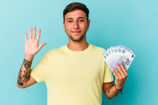 青い背景に孤立した紙幣を持つ若い白人男性笑顔陽気に数字を示します5とともに指 — ストック写真