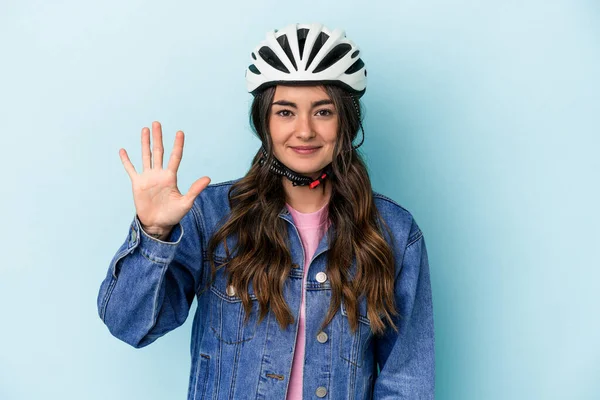 Young caucasian woman rinding a bike isolated on blue background smiling cheerful showing number five with fingers.