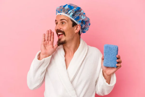 Young caucasian man going to the shower holding sponge isolated on pink background shouting and holding palm near opened mouth.