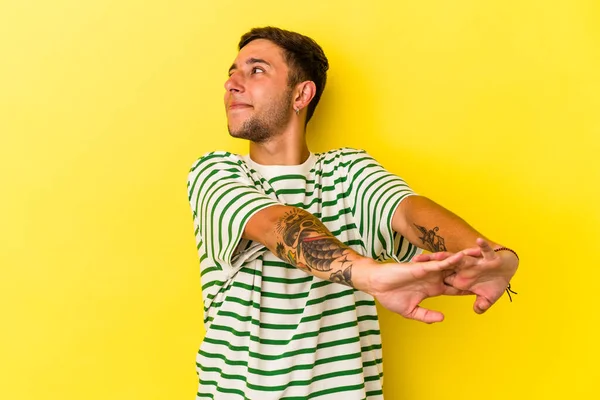 Sarı arka planda izole edilmiş, kollarını esneten, rahat bir pozisyonda dövmeleri olan genç beyaz bir adam..