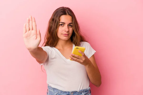 年轻的高加索女人用粉色背景隔离的手机站着 伸出的手显示了停止的标志 阻止了你 — 图库照片
