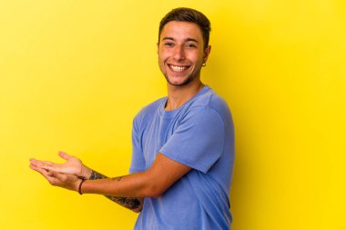 Sarı arka planda izole edilmiş dövmeleri olan genç beyaz bir adam palmiye ağacında bir kopya alanı tutuyor..