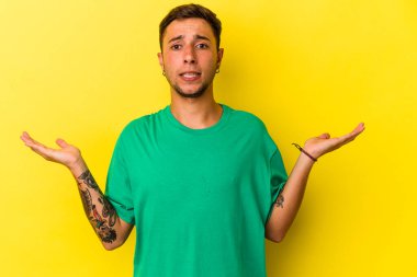 Sarı arka planda izole edilmiş beyaz dövmeli genç bir adam. Kafası karışmış ve omuz silkiyor..