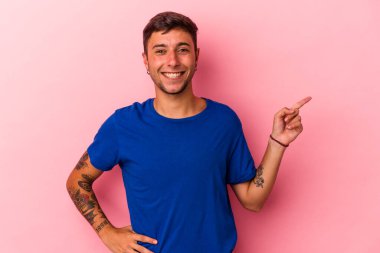 Sarı arka planda izole edilmiş dövmeleri olan genç beyaz adam işaret parmağıyla işaret ederek gülümsüyor..