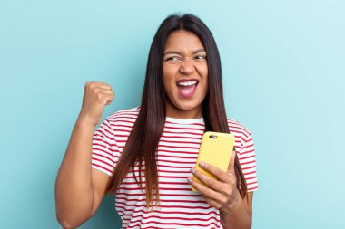 Elinde cep telefonu olan genç Venezuelalı kadın mavi arka planda izole bir şekilde zafer kazandıktan sonra yumruğunu kaldırıyor..
