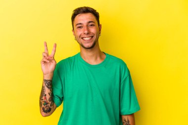 Sarı arka planda izole edilmiş beyaz dövmeli genç bir adam neşeli ve kaygısız parmaklarıyla barış sembolü gösteriyor..