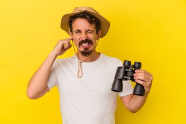 Dürbün tutan genç beyaz adam sarı arka planda izole edilmiş kulakları elleriyle kapatıyor..