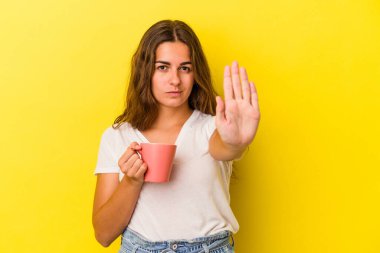 Sarı arka planda izole edilmiş bir kupa tutan genç beyaz kadın elini uzatmış, dur işareti göstererek seni engelliyor..