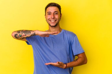 Sarı arka planda dövmeleri olan beyaz bir adam iki eliyle bir şey tutuyor, ürün sunumu..