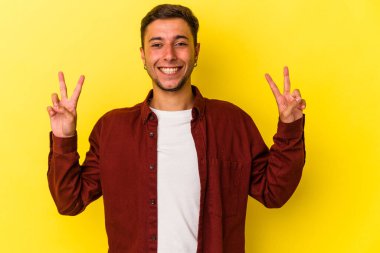 Sarı arka planda izole edilmiş dövmeleri olan genç beyaz adam zafer işareti gösteriyor ve gülümsüyor..