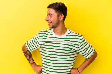 Sarı arka planda dövmeleri olan beyaz bir genç adam mutlu bir şekilde gülüyor ve ellerini karnının üstünde tutarak eğleniyor..