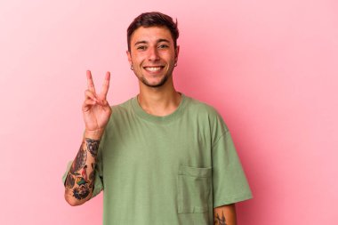 Sarı arka planda izole edilmiş dövmeleri olan genç beyaz adam zafer işareti gösteriyor ve gülümsüyor..