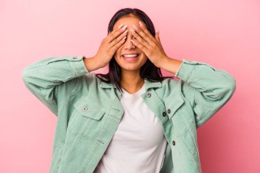 Pembe arka planda soyutlanmış genç Latin bir kadın elleriyle gözlerini kapatıyor, geniş bir gülümsemeyle bir sürpriz bekliyor..