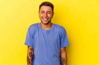 Sarı arka planda izole edilmiş dövmeleri olan genç beyaz adam gülüyor ve gözlerini kapıyor, rahatlamış ve mutlu hissediyor..