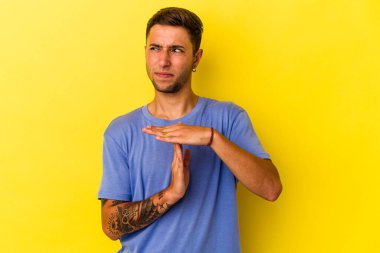 Sarı arka planda izole edilmiş dövmeleri olan genç beyaz bir adam, bir zaman aşımı hareketi gösteriyor..