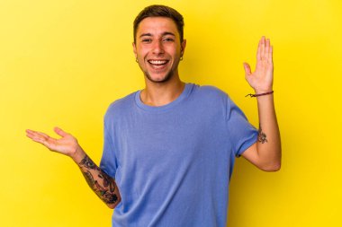 Sarı arka planda dövmeleri olan beyaz bir genç avucunun içinde fotokopi alanı tutuyor, el üstünde tutuyor. Şaşırmış ve mutlu..