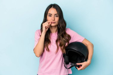 Young caucasian woman holding a motor bike helmet isolated on blue background with fingers on lips keeping a secret.