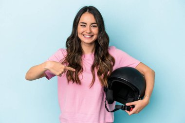 Young caucasian woman holding a motor bike helmet isolated on blue background person pointing by hand to a shirt copy space, proud and confident