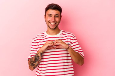 Sarı arka planda izole edilmiş dövmeleri olan genç beyaz bir adam gülümsüyor ve elleriyle kalp şeklini gösteriyor..