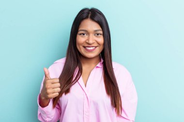 Genç Venezuelalı kadın mavi arka planda izole edilmiş gülümsüyor ve başparmağını kaldırıyor.