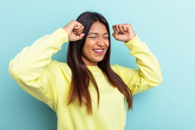 Genç Venezuelalı bir kadın mavi arka planda izole edilmiş özel bir günü kutluyor, atlıyor ve enerji ile kollarını kaldırıyor..