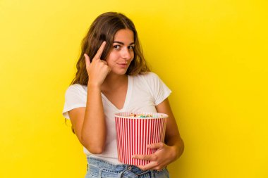 Sarı arka planda izole edilmiş patlamış mısır tutan beyaz bir kadın şakağı parmağıyla işaret ediyor, düşünüyor ve bir göreve odaklanıyor..