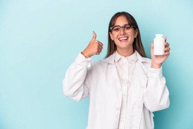 Genç eczacı kadın mavi arka planda izole haplar tutuyor gülümsüyor ve parmağını kaldırıyor.